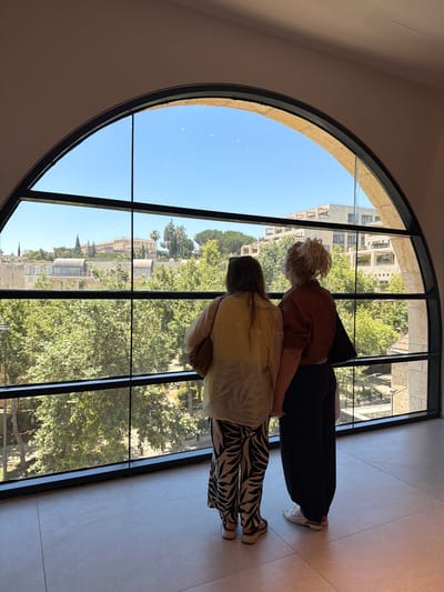 Two women stand looking out of a large arched window onto a cityscape of Jerusalem, Israel. The woman on the left wears a cream-colored top and black and white patterned pants, while the woman on the right wears a reddish-brown top and dark pants. They are positioned in the foreground, with their backs to the camera. Through the window, a clear blue sky is visible above buildings and lush green trees. The architecture suggests a modern or contemporary building with the large window. The setting appears to be a public space or a visitor center, given the vantage point.