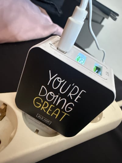 A close-up shot shows a white and black multi-port USB charger plugged into a white power strip. The charger has two illuminated USB ports, one showing a bright blue light and the other a greenish light. The charger has text on its black surface that reads "YOU'RE DOING GREAT" in white lettering, with "ALE-HOP" printed in a small black and white rectangle below it. A white USB cable is plugged into one of the ports. The background is blurred, but suggests a desk or table surface with dark and light-colored objects. The power strip appears to be a standard European model, suggesting the location is likely in Spain, specifically Alcalá De Henares. The lighting suggests an indoor setting, possibly during the day given the brightness of the illuminated ports. The overall impression is of a functional item in use.