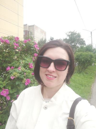 A mid-shot, selfie-style photograph shows a woman in her late 30s with dark brown, shoulder-length hair and wearing large, dark-rimmed sunglasses and a white jacket. She has on bright pink lipstick and is smiling. She is standing in front of a large shrub with pink flowers and green leaves, with a light-colored building to her left and a paved walkway to her right. In the background, there are more trees and buildings, consistent with an urban setting in Tosno, Russia. The sky is overcast.