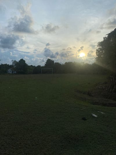 The image depicts an outdoor scene on a grassy field, possibly a sports field, with a soccer goal visible in the distance. The sun is shining brightly through a cloudy sky, suggesting it is daytime. The overall lighting indicates it could be late afternoon or early morning, with the sun casting a warm glow.
To the left of the frame, the sky is filled with dramatic clouds, some dark and heavy, others wispy and illuminated by the sun. Silhouetted trees frame the upper left edge of the image. To the right, the grassy field stretches out, appearing well-maintained. In the mid-ground, a line of trees separates the field from the sky, and a white building with a tiled roof is partially visible amongst them, suggesting a residential area nearby.
On the grassy area in the foreground, there are a few scattered objects, including what appears to be a plastic bottle and a dark piece of trash. These details suggest the area may not be entirely free of litter.
The scene is devoid of people, giving it a tranquil or possibly abandoned feel. The presence of the soccer goal implies the field is used for sports, but the lack of activity makes it hard to determine if an event is currently taking place. The natural elements, like the sun and clouds, create a somewhat serene atmosphere, juxtaposed with the man-made elements of the soccer goal and the distant building. The location context suggests this is Keudeublang, Indonesia. There is no visible text in the image.