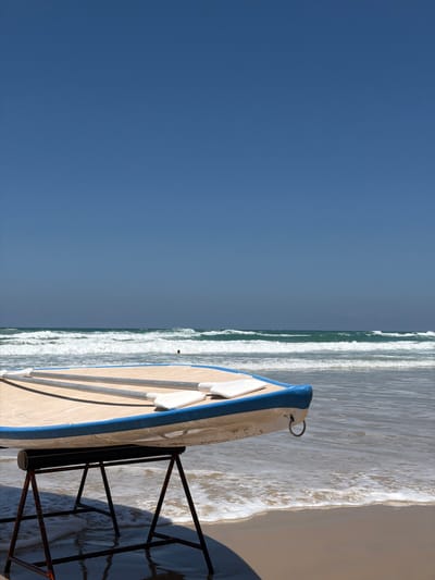 A high-angle shot depicts a partly visible beach scene in Bat Yam, Israel, focusing on a boat and its surrounding environment. The left side of the frame is dominated by a clear blue sky. The right side reveals the ocean with small, foamy waves breaking onto the sandy beach. A small boat, painted white with a blue trim, is stationed on the sand, partially submerged in the shallow water. A dark, rusted metal boat stand sits under the boat, partially submerged in the water. The image suggests a sunny day at the beach.