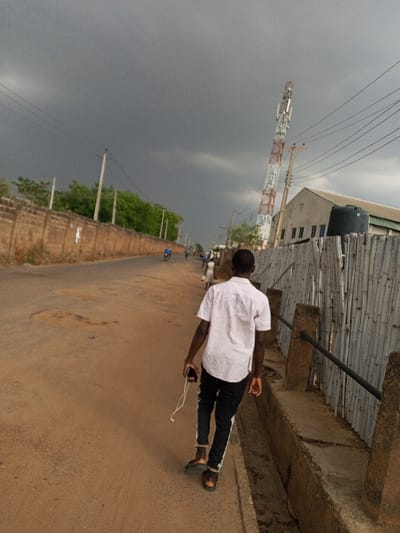 The image depicts an outdoor street scene in Ilorin, Nigeria, under a heavily overcast, dark grey sky. A young male, viewed from behind, walks away from the camera on the right side of a dirt road. He wears a light pink short-sleeved collared shirt, black trousers with white side stripes, and dark sandals, holding a black smartphone with white wired earbuds in his left hand. To his immediate right, a shallow concrete drainage ditch runs parallel to the road, separated by concrete posts and a dark horizontal metal bar. Beyond this, a fence made of vertical wooden slats or bamboo panels lines the roadside, partially obscuring a light-colored industrial-style building with a dark water tank on its roof. A tall, red and white striped telecommunications tower stands prominently adjacent to the building. On the left side of the road, a long, unpainted brick wall with green foliage behind it extends into the distance. Several utility poles with power lines run along this wall. Further down the road, multiple distant figures, including individuals on motorcycles, are visible.
