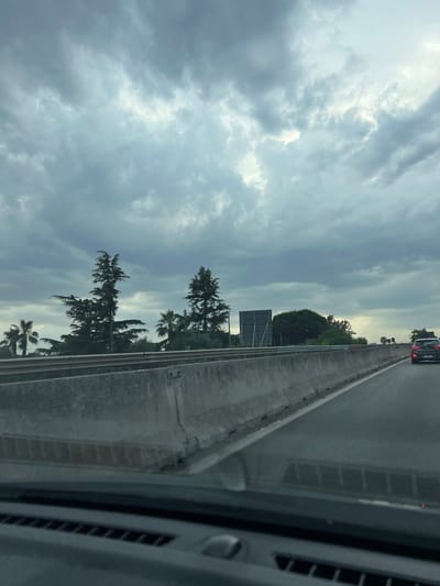 The image captures a view from a vehicle traveling on a highway. A concrete barrier separates lanes of traffic, with a dark-colored car visible ahead on the right. The sky is overcast with dramatic, cloudy formations. To the left of the highway, a line of trees and palm trees are visible, with a large, dark billboard-like structure positioned between them. The foreground shows the interior of the vehicle, including the dashboard. The scene is devoid of any specific urban elements that would pinpoint it to Bisceglie, Italy, other than the context provided.