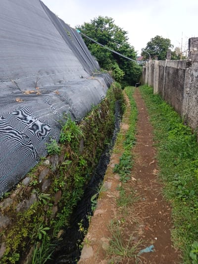 The scene, located in Koipa, Papua New Guinea, depicts a narrow, unpaved dirt pathway extending into the background under an overcast sky. To the immediate left, an elevated, sloped surface is covered entirely with black, woven mesh fabric exhibiting visible horizontal striations. Below this fabric, a stone retaining wall, heavily overgrown with moss and small green plants, borders a dark, narrow drainage channel containing water. To the right, a weathered concrete wall, constructed from vertically segmented panels, runs parallel to the path. The pathway itself is reddish-brown earth with patches of green grass and weeds along its edges. A small, light blue piece of plastic debris is visible on the path in the lower right foreground. Beyond the visible structures in the background, dense green foliage and trees are present. No individuals, animals, or discernible actions are depicted.
