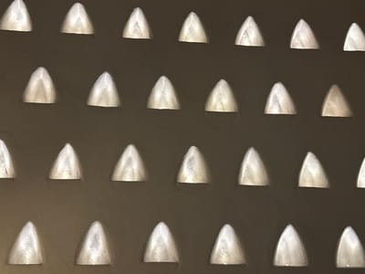 A pattern of triangular recessed lights is set in a dark brown or gray wall. The lights are arranged in a grid. The interior of each triangle is lit with a soft, white glow, creating a three-dimensional effect. The wall's surface appears textured, possibly stucco or a similar material. While the prompt mentions Ramat Negev Regional Council, Israel, there is no explicit evidence in the image to confirm this location.