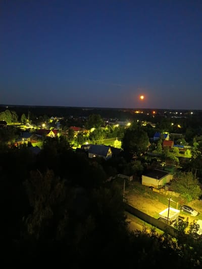 This is an aerial, nighttime photograph of a residential area in the city of Alexandrov, Russia. The image shows a cluster of houses with lit windows, streetlights, and a bright, partially obscured moon or sun low on the horizon, casting a reddish-orange glow. The landscape is characterized by dense foliage and trees, with some structures visible amidst the greenery. The foreground features dark, silhouetted trees, suggesting the photograph was taken from a slightly elevated position. The scene depicts a quiet, rural night in a Russian town.