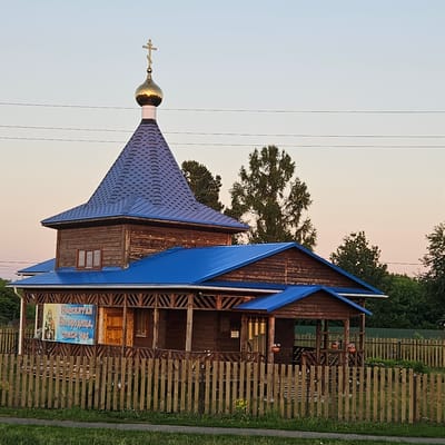 The image shows a small wooden Orthodox church in the village of Novostroyka, Russia. The church has a blue metal roof and a golden dome with a cross on top. There is a wooden fence in front of the church, and a banner with an image of a religious icon and text in Cyrillic is hanging on the side of the building. The sky is clear and the sun is setting, casting a warm glow on the scene. Trees and greenery surround the church, indicating a rural setting.