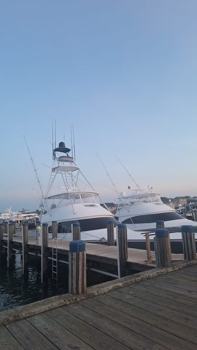 This is a medium-angle, vertical, outdoor, eye-level photograph of two white yachts docked side-by-side at a wooden pier. The sky is a clear, pale blue with no clouds. The yachts are large, white vessels with dark trim along the hull. Both have tall, silver towers with multiple outriggers, suggesting they are fishing boats. The boat on the left is closer to the camera and appears to be slightly larger. The pier is made of weathered wooden planks and is supported by thick, wooden pilings topped with blue caps. The water around the pier is dark and still. In the background, beyond the yachts, there is a cluster of houses and more boats docked. The scene appears to be at dusk or dawn. The image was taken in Nantucket, United States of America.