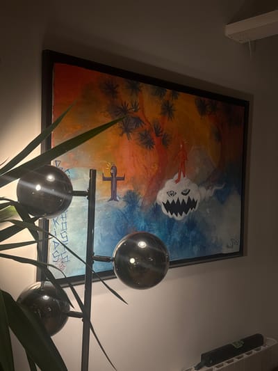 The image displays an indoor scene in Saint-Denis, France, centered on a large, framed, abstract painting mounted on a light-colored wall. The painting features a vibrant background, transitioning from warm hues of red, orange, and yellow in the upper section to cooler blues and purples below. Dark, stylized tree-like forms with star-shaped leaves are depicted in the upper portion. The artwork includes two humanoid figures: a purple figure with outstretched arms, an oval head, and a yellow circular symbol containing a white 'G' positioned above it, seen on the left. Faint, vertical text is partially visible behind this figure. On the right, a red humanoid figure with an oval head stands on a white, cloud-like entity. This entity exhibits two distinct eyes—one with a spiral pupil and the other with a circular pupil—and a prominent mouth with jagged, sharp teeth. A small, illegible signature is present in the painting's bottom right corner.

In the immediate foreground on the left, a dark metallic floor lamp with three spherical, reflective lamp heads is partially obscured by the long, green leaves of an indoor plant. Reflections of interior light sources are visible on the lamp heads. To the right, at the bottom of the frame, a white radiator is visible, upon which rests a dark, cylindrical object. A white architectural beam extends from the wall in the upper right quadrant of the image. The scene is illuminated by ambient interior lighting.
