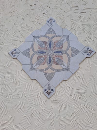 The image depicts a decorative tile arrangement mounted on a textured, light-beige wall. The tile design is symmetrical and floral, composed of multiple smaller tiles with a central rosette pattern. The rosette features geometric shapes in shades of blue, beige, and gray. The textured wall background shows an irregular pattern of thick, short strokes, resembling stucco or plaster. There is no information available in the image to suggest the location is Elektrostal, Russia. The scene appears to be a close-up interior shot.
