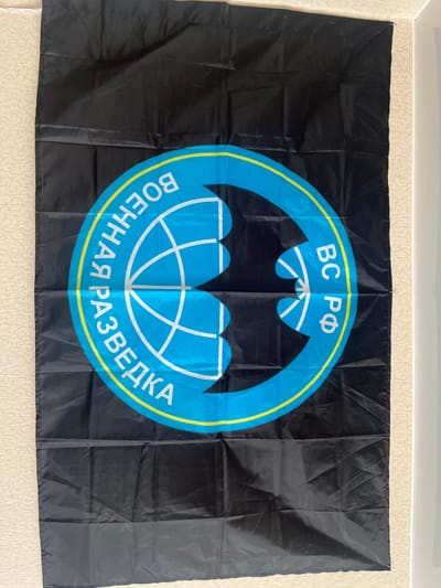 A black flag with a circular emblem is displayed vertically against a textured wall. The emblem features a blue circle outlined in yellow, with a stylized globe or network design inside. White text in a Cyrillic alphabet encircles the globe. On the left side of the circle, the text reads "ВОЕННАЯ РАЗВЕДКА" (Military Intelligence). On the right side, it reads "ВС РФ" (Armed Forces of the Russian Federation). The flag appears to be made of a synthetic fabric and is creased in several places, suggesting it has been folded or stored. The background wall has a subtle, repeating pattern. The lighting is even, with no strong shadows, indicating ambient light conditions. No people or any other objects are visible in the scene. The image is taken from a slightly elevated, straight-on perspective.