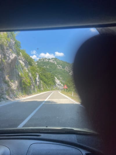 A forward-facing, eye-level shot captures a paved mountain road in Dragalj, Montenegro, on a clear, sunny day. The road curves gently to the right, with a white double line painted down its center, indicating passing is permitted. The terrain is mountainous, with steep, rocky inclines covered in lush green vegetation on the left side of the road. A metal mesh fence runs along the mountainside, presumably to prevent rockfalls. On the right side of the road, a red triangular warning sign with a white border is visible. In the distance, more mountains covered in trees can be seen under a bright blue sky with scattered white clouds. The lower part of the image shows the dashboard and windshield of a vehicle, with a dark, blurry object obscuring the right side of the view. The scene appears to be taken from inside a moving vehicle.