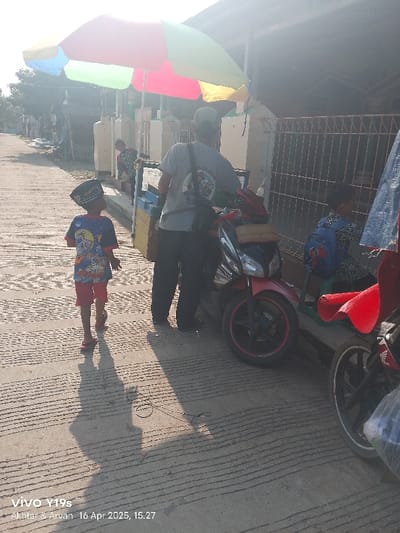 The scene is a street in Anjatan, Indonesia (latitude: -6.419, longitude: 107.979). A man in a gray shirt is standing near a parked black motorcycle, which is in the middle of the street. A young boy with a black hat and a blue and red shirt is walking in the opposite direction. A second man is seated behind a red blanket on the sidewalk, which is lined with a metal fence. A young boy in a blue shirt is seated next to the red blanket. A colorful umbrella is positioned to the left of the scene. The ground is paved with concrete and has a textured surface. The scene is lit with a bright, sunny light.  The photograph was taken at 15:27 on April 16, 2025.
