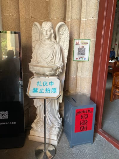 This vertical shot, rotated 90 degrees clockwise, captures an indoor scene likely within a church or cathedral, specifically identified as the Guangzhou Sacred Heart Cathedral.

In the foreground, a large white statue of an angel with detailed wings stands, holding an object in its arms. Attached to the angel's base is a white sign with blue Chinese characters stating, "礼仪中禁止拍照" (Lǐyí zhōng jìnzhǐ pāizhào), meaning "No photography during ceremony/ritual."

To the right of the angel statue, a grey "Offering Box" is visible. It features red labels with white text that read "Offering Box" in English and "奉獻箱" (Fèngxiàn xiāng), meaning "Offering Box," in Chinese.

On the stone wall behind the angel, a green sign with a QR code and Chinese text is displayed. The text includes a "温馨提示" (Wēnxīn Tíshì - Warm reminder) advising visitors to "建议参观圣心大教堂 请勿大声喧哗" (Jiànyì cānguān shèngxīn dà jiàotáng qǐng wù dàshēng xuānhuá), which translates to "When visiting the Sacred Heart Cathedral, please do not make loud noises." Below the QR code, "广州石室圣心大教堂" (Guǎngzhōu Shíshì Shèngxīn Dàjiàotáng), or "Guangzhou Sacred Heart Cathedral," is written, along with contact numbers (e.g., 020-83336711) and social media icons.

In the bottom left, a reflection in a dark, possibly glass, surface shows a partial view of what appears to be a person and outdoor greenery. Another QR code and the text "聖心大教堂" (Shèngxīn Dàjiàotáng - Sacred Heart Cathedral) are faintly visible on this surface. In the background, wooden pews and a person are partially visible, reinforcing the church setting. The lighting is bright, suggesting daytime.