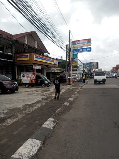 The scene depicted is an urban streetscape. This location is identified as likely being in Indonesia based on prominent "Indomaret" convenience store signage and multiple Indonesian bank logos (BCA, Mandiri, BSI). The provided location of Gubbisamamidi, India, is not supported by visual evidence.
The image captures a daytime view under an overcast sky. In the foreground, a worn dark grey paver sidewalk with a faded white curb line separates the viewer's position from an asphalt road. A male subject, wearing a dark hooded long-sleeve shirt with white graphics on the back and dark trousers, walks away from the camera on this sidewalk.
To the immediate left of the sidewalk is a paved parking area. A maroon passenger car with a license plate (F 1689 KKY) is parked. Adjacent to it, a black delivery truck with a white refrigerated unit, branded "Cleo," is also parked.
Behind the paved area stands a two-story building. The ground floor houses an "Indomaret" convenience store, identifiable by its prominent blue, red, and yellow striped sign indicating "Indomaret 24 JAM" (24 Hours). The upper floor features a brown-tiled gable roof and visible balconies.
In the mid-ground, a tall black utility pole rises from the sidewalk, heavily laden with numerous black electrical cables crisscrossing the upper frame. Attached to this pole are multiple vertical signs, including another "Indomaret 24 JAM" sign, a "PAKET" sign, and various service icons (ATM, food, car). Below these are distinct signs for "ATM BNI," "ATM BCA," "ATM Mandiri," "POSPAY," "Mazda," and "BSI."
On the asphalt road, a white minivan is observed heading away from the viewer. Near the utility pole, another individual in a light-colored uniform stands. The road recedes into the background, showing additional vehicles, buildings, and multiple advertising billboards on the right side of the street.