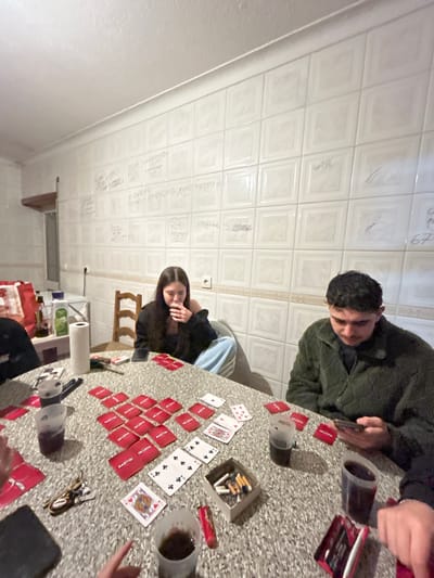 The image depicts an indoor, informal social gathering in Bragança, Portugal, characterized by its distinctive tiled walls. These off-white, square ceramic tiles are extensively covered with handwritten inscriptions, signatures, and simple drawings in various dark markers, primarily in Portuguese (e.g., "O DINIS COMEU SAPUCAIAS," "67"). This style of inscribed walls is characteristic of informal taverns or student gathering places in the city. The ceiling is plain and textured white.
In the foreground, a speckled light-grey table is the central point of activity, hosting a card game involving at least four individuals. The game setup includes numerous red-backed "MARTINI" branded cards, some face down, others forming stacks. Several standard playing cards, such as the Ace of Spades, 4 of Spades, 8 of Clubs, 10 of Spades, 9 of Clubs, and Queen of Hearts, are visible face-up.
Multiple clear plastic cups containing a dark liquid are distributed around the table. An improvised ashtray, a small cardboard box, holds cigarette butts and loose tobacco/cigarettes, with a red lighter nearby. Personal items include a set of keys with a keychain, and at least two smartphones; one rests on the table in front of a female subject, and another is actively held by a male subject. A dark, rectangular object, possibly another smartphone or tablet, is partially visible in the bottom left. A roll of white paper towels is also on the table.
Of the people present, a female individual with long, dark hair is seated center-left, wearing a dark top and light blue pants. She is seen bringing a clear plastic cup to her lips. To her right, a male individual with dark, curly hair is seated, wearing a dark green textured jacket over a dark shirt. He is looking down at a smartphone held in both hands. Portions of other individuals are visible: a dark sleeve and a leg in light blue pants are on the far left, and a hand holding a cup is in the bottom right.
In the background to the left, a surface holds a dark red bag, multiple bottles (one dark-colored, possibly olive oil, and another containing a light green liquid), and a white roll of paper towels. A light-colored wooden chair is also visible. The overall scene suggests a casual, late-night social interaction among friends or acquaintances.