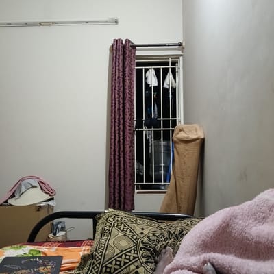 The interior shot depicts a room, potentially a bedroom, in Bengaluru, India, suggested by the presence of Indian-style curtains and bedding. The room is sparsely furnished, with a bed dominating the foreground and a window to the left. The bed is covered with patterned sheets and pillows and topped with a pink blanket, partially obscuring a person lying in it, with only a foot visible. Beside the bed to the left, there is a cardboard box topped with clothing. The window is fitted with white bars and covered with curtains on the left. Outside the window, clothes are hanging. Against the wall to the right of the window rests a rolled-up mattress. The walls are painted a muted white, and a fluorescent light fixture is visible on the ceiling.