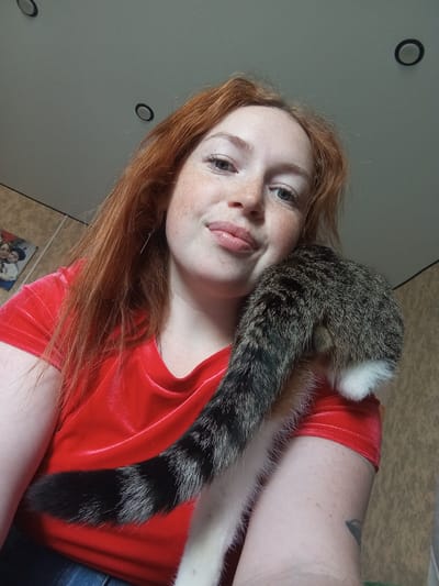 A pale-skinned, reddish-haired woman with freckles is holding a cat. She is wearing a red velvet top. The cat is a tabby with a white-tipped tail. The background includes a beige wall with circular light fixtures. The photo was taken indoors, and it is hard to tell if it was taken in Yurgamysh, Russia or any other specific place.