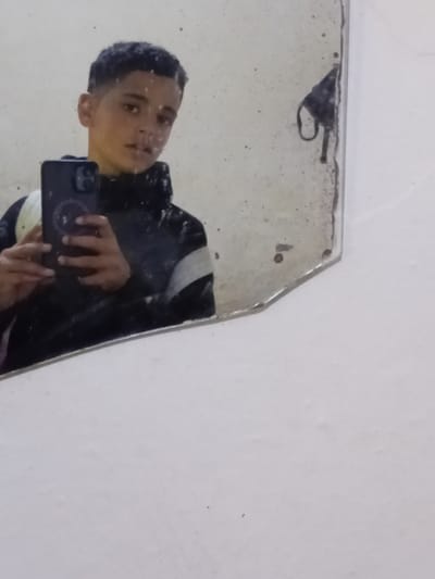 An indoor scene captured through a mirror reflection shows a young person taking a selfie. The person, a young male, is seen from the chest up, reflected horizontally in an old, somewhat dirty mirror. He has short, dark curly hair and is looking directly towards the camera of the smartphone he is holding. He is wearing a dark jacket with a white stripe over what appears to be a lighter-colored shirt. His right hand holds the dark-cased smartphone (possibly an iPhone due to the distinct camera bump design) up, angled towards himself to capture the reflection.
The mirror itself is rectangular but features an irregular, possibly chipped or crudely cut, upper left corner. Its surface is noticeably dusty and smudged, with various small dark spots suggesting wear and tear or accumulated dirt. In the very upper left corner of the mirror, a dark object resembling a folded face mask is visible, seemingly stuck to the mirror or resting on its edge. The background outside the mirror's reflective area is a plain, off-white wall, which also shows some minor imperfections or texture. The overall setting appears to be a casual indoor space, possibly a room or a bathroom, in Martil ⵎⴰⵔⵜⵉⵍ مرتيل, Morocco. No visible text is present in the image.
