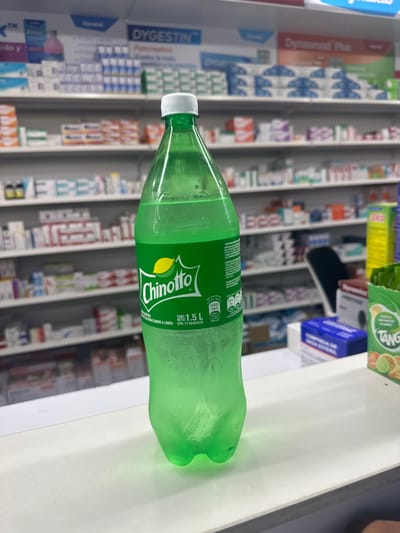 A 1.5L bottle of Chinotto lemon-flavored soda lies horizontally on a white surface in what appears to be a pharmacy in Parroquia Ocumare, Venezuela. Rows of pharmaceuticals in boxes and packages fill shelves in the background, including Dynasmed Plus and Dygestin. Additional products, including Tang flavored drink mix and "Dynamics Prestige" brand items, are partially visible on the surface near the bottle. The Chinotto bottle is green with a white cap.