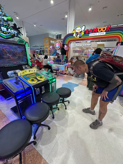 This image captures a scene inside an arcade, likely in Wichit, Thailand, given the location context. The main subject is a man with tattoos on his arms and neck, wearing a black t-shirt and blue shorts, with a backpack on. He is bending down, looking at his phone. Several arcade machines are visible, including a "Blast" game and a "Gunslinger" game with blue LED lighting. Other people, including children, are present in the background, some appearing to be playing games or walking through the arcade.

The arcade is brightly lit, suggesting it's daytime or a well-lit indoor space. A prominent sign reads "SANOOK GAMES ZONE" in English and Thai, with a cartoon bunny character. Another sign says "Stamp Corner." The floor is tiled with a speckled pattern. The overall atmosphere appears to be casual and recreational, typical of an entertainment venue. The presence of arcade games, colorful signage, and people enjoying themselves points to a lively and engaging environment.