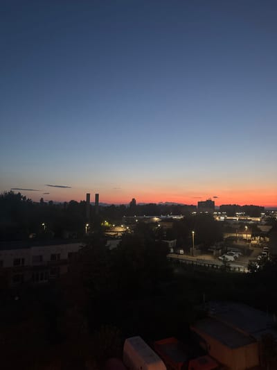 This is an outdoor, high-angle shot of a city skyline at dusk. The sky transitions from a deep blue at the top to a vibrant orange and red near the horizon. In the mid-ground, there are silhouetted trees and various buildings, including what appear to be factories or industrial structures with tall chimneys. Streetlights illuminate some areas of the city. In the foreground, there are darker, silhouetted trees and some objects that appear to be vehicles or containers. The overall scene suggests an urban industrial area at the end of the day. While the specific location within Pazardzhik, Bulgaria is not identifiable from the visual information alone, the image depicts a common urban twilight scene. There are no visible people or specific interactions.