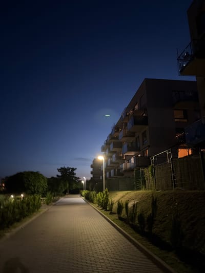 The image depicts a paved walkway lined with manicured bushes, leading towards a modern apartment building on the right. The sky is dark blue, suggesting it is either dusk or night. Streetlights illuminate the walkway, casting long shadows from the bushes on the left and the building's facade on the right. The apartment building has multiple balconies with glass railings, and some lights are visible inside the windows. A fence runs along the base of the building. The overall scene is residential and quiet. This image likely captures a view in the Bystrzyca district of Lublin, Poland, given the architectural style and the surrounding greenery often found in newer developments there.