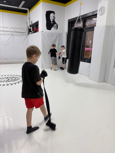 This image depicts an indoor martial arts or combat sports training facility in Novosaratovka, Russia. The room features white padded walls extending from a white mat floor, which has a black decorative pattern in the foreground. The upper walls are white, transitioning to a black ceiling with a yellow border. A large black and white decal of an elderly Asian man's portrait is affixed to a corner wall.
In the foreground, a young boy with light brown hair, dressed in a black t-shirt and red shorts, is in motion, unwrapping a long black fabric strip, likely a hand wrap. He wears black foot coverings.
In the midground, towards the right, two other young boys are preparing. One boy, wearing a black t-shirt and camouflage-patterned shorts, has purple protective gloves on his hands. The second boy, wearing a white t-shirt with black detailing and black shorts with a white stripe and the number "7," is assisting the first boy with his hand wraps or gloves. Both boys are wearing socks.
A black cylindrical punching bag hangs from the ceiling by chains in the midground, positioned to the right of the two boys. A window behind the punching bag shows reflections and internal signage with what appears to be Russian text, including "АННА," and a stylized figure. A white analog clock is visible on the upper right wall. The facility is illuminated by overhead lighting, and equipment or bags are visible on a shelf or bench along the left wall.