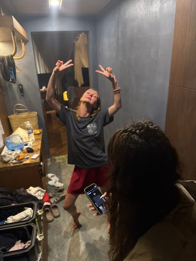 This indoor scene, set in Mù Cang Chải, Vietnam, features two individuals within a room characterized by gray, textured walls and dark floor tiling.

In the mid-ground, a light-skinned female with light brown hair and wearing a dark gray t-shirt and red shorts, stands barefoot. Her head is tilted back with eyes closed, and both arms are raised, hands forming peace signs. Colorful bracelets adorn her right wrist, and a ring is visible on her left index finger.

In the foreground, partially visible from the back, a person with dark, braided hair and wearing a light brown garment holds a light green smartphone, pointed towards the first individual. The phone's screen displays a dark interface.

The room's left side features a wooden wall-mounted shelf with hangers, from which dark clothing hangs. Below this, a wooden counter is cluttered with a woven straw basket, a partially full plastic water bottle, yellow snack packaging, folded clothing, a blue plastic bag, a black remote control, and a small white object. On the floor below the counter, two open black luggage cases contain various garments. Additional footwear, including white slippers, brown slip-on shoes, and dark sandals, is visible on the floor, alongside a red Santa hat. A bright ceiling light fixture is centered at the top of the image. Through a doorway or opening in the background, a window with light-colored curtains and another ambient light source are visible.