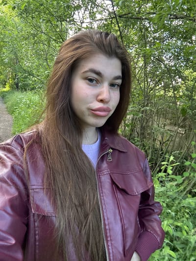 A young woman with long, brown hair and a maroon faux leather jacket poses for a selfie outdoors. She has light skin with freckles and is wearing a light purple t-shirt underneath her jacket. Her lips are slightly parted and she is looking towards the camera. The background consists of lush green trees and foliage, suggesting a natural, possibly rural or park-like setting. The path to her left indicates a walking trail. The location is identified as Мещёры, Russia.