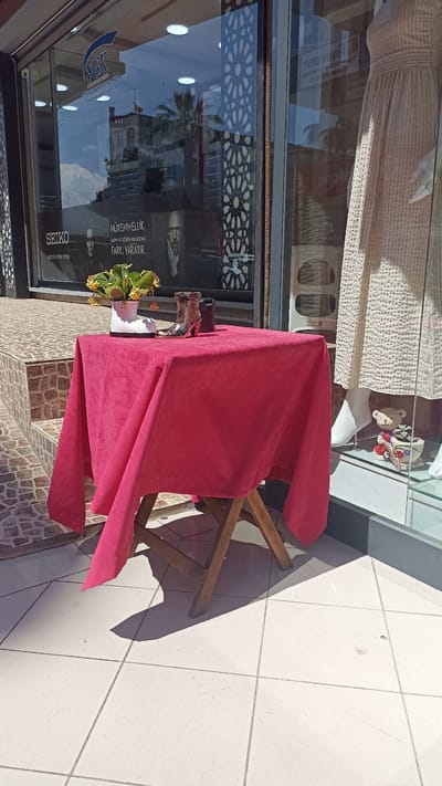 A small, circular, wooden table draped with a bright pink tablecloth is placed on a tiled outdoor surface in front of a storefront. On the table, a white ceramic boot holds a small plant with yellow flowers, accompanied by a metallic boot-shaped object and a dark cylinder. The storefront window behind the table reflects the sky, clouds, and buildings, and displays advertisements for "SGK" and "SEIKO." To the right of the table, a mannequin dressed in a cream-colored, textured garment stands within the store, visible through the glass. The scene appears to be located in Yenişehir, Turkey.