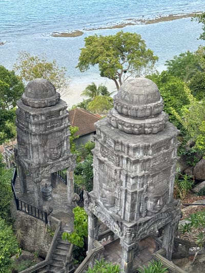 The image captures an aerial view of two ornate stone structures, likely part of a temple or memorial, situated on a hillside overlooking a body of water and a beach. These structures feature intricate carvings, including human-like faces and decorative motifs. Lush green foliage surrounds the structures and a path with stone steps leads down from them. In the background, the turquoise ocean stretches towards the horizon, with a sandy beach visible in the distance. A few buildings with reddish-brown roofs are also scattered among the trees, suggesting a settled area nearby. The scene conveys a sense of ancient artistry set against a tropical coastal landscape. The specific location is identified as Ban Tai Subdistrict, Thailand.
