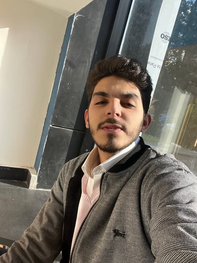 A single male individual, appearing to be between 18 and 25 years old, is depicted in a self-portrait. He has dark brown hair, dark eyes, and a trimmed dark beard and mustache. He is wearing a white collared shirt, a light pink V-neck sweater, and a grey and black houndstooth pattern bomber jacket with a black ribbed collar and a black embroidered animal logo on the left chest. The individual is looking directly into the camera with a neutral expression.

The background consists of an indoor environment with significant natural lighting. To the subject's left, a plain off-white wall meets a dark grey or black vertical structural column. To the right, a large glass window or panel is present, reflecting bright sunlight and revealing blurred outdoor elements, including greenery. A white label is affixed to the glass, displaying partial text including "ISO" within a circular logo, "white Pane," and "er installation." The specific location within Al-Mazza Municipality, Syria, is not identifiable from the visual evidence provided.