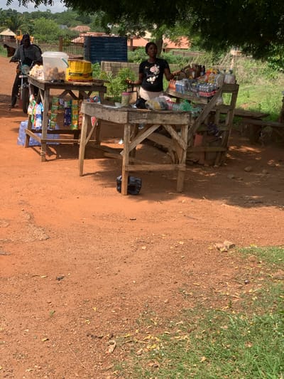 A street scene in Kabba, Nigeria shows a dirt road with a wooden roadside stand. The stand is stocked with packaged food items and beverages. A woman stands behind the stand. In the background, there are trees and a building. Another person sits on a parked motorcycle near the stand.