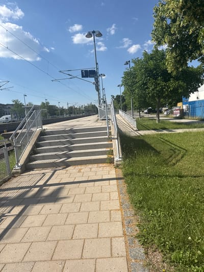 The image depicts the exterior of the Höhenkirchen-Siegertsbrunn train station in Germany on a clear, sunny day. In the foreground, a paved walkway made of light-colored rectangular stones leads towards a set of concrete stairs. The stairs are bordered by metal railings and are partially in shadow. To the right of the stairs, a grassy area with some small yellow flowers is visible. Beyond the stairs, the train platform is visible, with more paved areas and grassy sections. Overhead, train catenary wires and lampposts are strung across the sky. In the background, trees line the edge of the station area, and beyond them, buildings and parked vehicles can be seen. A blue and white building with some signage is also present in the distance. The overall scene is that of a functional, well-maintained public transportation hub in a suburban or semi-urban setting.