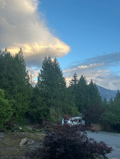 The image captures a landscape in Squamish, Canada. The sky exhibits a mix of blue and cumulus clouds, some illuminated by sunlight. In the background, a mountain peak is visible. The foreground is dominated by trees of various heights and species, including dark-leaved Japanese maples. A white RV is parked near a wooden structure. The ground appears rocky and vegetated.