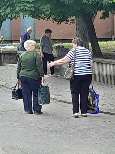 Two women and a man are standing on a paved area with trees and buildings in the background. The woman on the left is wearing a green sweater and dark pants, carrying a black handbag in her left hand and a dark green bag in her right hand. The woman on the right is wearing a blue and white striped shirt and dark pants, carrying a patterned handbag on her left shoulder and a blue shopping bag in her right hand. She is pointing her right hand towards the left. The man, in the background, is wearing a grey shirt and dark pants, with his right hand to his face. Another person, wearing a dark purple top and dark pants, is partially visible behind the woman on the left. The ground is paved with cobblestones. The environment appears to be an outdoor public space, possibly in the city of Berezhany, Ukraine, given the architectural style of the background buildings.