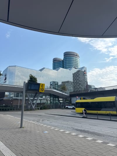 The image shows a modern urban scene in Utrecht, Netherlands, on a clear, sunny day. In the foreground, a gray canopy extends from the left edge of the frame, casting a shadow. To the right, a paved pedestrian walkway with a white-lined curb runs alongside a road. A yellow bus is parked on the side of the road, with its doors closed. A white car is also visible nearby.
Dominating the background are several contemporary glass-facade buildings. One prominent building features a curved, cylindrical design with glass panels. Another building has a geometric, angular structure, also covered in glass. The buildings appear to be office complexes or commercial spaces, possibly a business district. The sky above is blue with scattered white clouds.
A yellow sign with black text "C 10" is mounted on a metal pole in front of the buildings. Below this sign is a digital display showing a schedule or information, with text too small to read clearly. The overall impression is one of a bustling, modern city environment. The lighting suggests it is daytime, likely late morning or early afternoon. There are no discernible people engaged in activity, but the presence of the bus and cars indicates ongoing movement and activity within the urban setting. Some text is visible on the yellow bus, and the word "Rabobank" is partially visible on one of the cylindrical buildings.