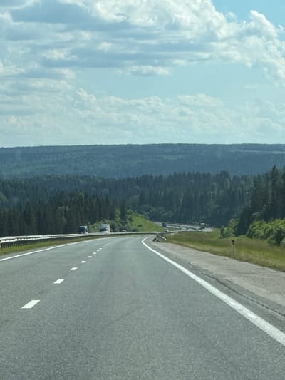 The image shows a road curving through a wooded landscape. The road is asphalt, with white markings indicating lanes and the edge. On the right side of the road, there is a grassy embankment leading up to a dense forest. The forest is primarily composed of dark green evergreen trees, with some lighter green deciduous trees mixed in at higher elevations. In the distance, there are rolling hills covered in trees, fading into a hazy blue.

The sky on the left side of the image is a bright blue, with large, fluffy white clouds. The lighting suggests it is daytime, likely late morning or early afternoon, with bright sunlight casting shadows. The weather appears to be clear and fair.

Several vehicles are visible on the road in the middle ground, appearing to be trucks or vans. They are following the curve of the road and seem to be moving away from the viewer. There is a guardrail along the side of the road, separating it from the embankment and forest.

The overall scene evokes a sense of travel and journey through a natural, rural environment. The vastness of the forest and sky suggests a remote location. The image captures a serene and scenic moment, likely from a moving vehicle, given the perspective. There is no visible text in the image.