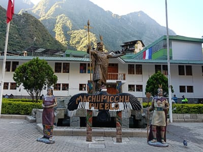 The image shows a plaza in the town of Machupicchu, Peru, located at the base of the Andes Mountains. In the foreground, from left to right, is a statue of a female figure, a sign reading "Machupicchu Pueblo" beneath a condor effigy, and a statue of a male figure. Behind these figures, there is a building painted white and green, with a Peruvian flag hung from a balcony. Behind the building are the mountains. The plaza floor is paved with light-colored bricks. There are people visible on a bench near a bush.