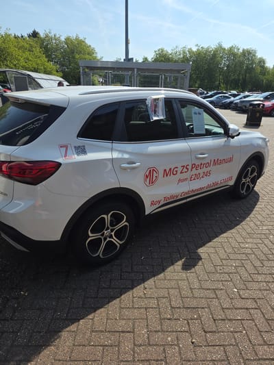 The image shows a white MG ZS Petrol Manual car parked on a paved surface, possibly at a car dealership. The car has "MG ZS Petrol Manual from £20,245" and "Roy Tolley Colchester 01206 574488" printed on its side, indicating that it is being sold at the Roy Tolley dealership in Colchester, United Kingdom. A "7 YEAR WARRANTY" sticker and QR code are visible near the car's rear light. Other cars are parked in the background, and trees are visible in the distance. The image is taken on a sunny day, as indicated by the shadow cast by the car.