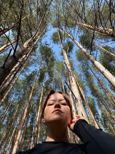 In an outdoor, upward-angled shot taken in a pine forest in Chaikovsky, Russia, a young woman with brown hair and makeup poses. She is wearing a black top and has her right hand raised to her ear. The background is dominated by tall, slender pine trees with clear blue sky visible between their branches. The perspective is from low down, looking up at the trees and the woman's face, which is partially lit by sunlight.