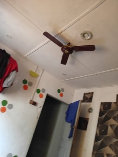 The image depicts an interior scene, likely a room. Dominating the upper portion of the frame is a ceiling fan with dark brown blades, partially in motion. Below the fan, on the white ceiling, a circular light fixture is visible.
The walls are painted white and are adorned with colorful circular decals, primarily in shades of green, orange, and grey. A yellow object, possibly a sponge, is attached to the wall to the left, near some hanging clothes that include a red garment and a grey undershirt.
To the right, a doorway or entrance leads into a darker area. Part of a patterned wall covering with a geometric design is visible on the right side of the frame. A blue piece of fabric, possibly clothing, is draped over something within the scene. The overall impression is of a living space.
The location provided for this image is Ipota, Nigeria.