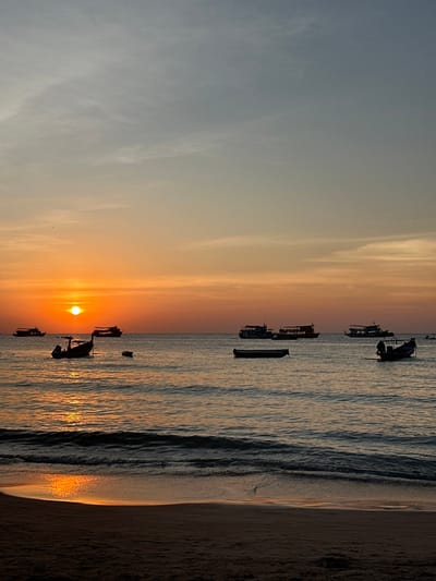 The image captures a sunset scene over a coastal area in Ko Tao, Thailand. The sun, a bright orange-yellow disc, is positioned low on the horizon, slightly left of the image's vertical center. Its intense light creates a luminous reflection path stretching across the calm ocean towards the foreground.
The sky exhibits a gradient from a light blue-grey at the top to vivid orange and yellow tones near the horizon. Wispy cloud formations are present in the upper and mid-sections of the sky.
The ocean surface is predominantly calm, with small, gentle waves visible as they approach the sandy beach in the foreground. The beach itself consists of dark, wet sand, occupying the bottom portion of the frame.
Six distinct vessels are present on the water, silhouetted against the bright sky and water reflections. In the midground, a long-tail boat is anchored left of center. To its immediate right, a smaller, dark, rectangular dinghy or rowboat is also anchored. Further to the right, another long-tail boat, appearing larger, is visible. In the background, four larger vessels, consistent with ferries or tour boats, are anchored: two are positioned on the left side of the frame, partially behind the sun, and two are on the right side, further out at sea. No individuals are visible in the scene.