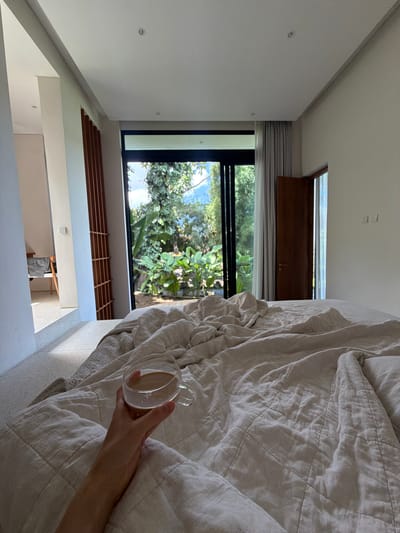 A vertical indoor shot shows a person's arm holding a clear glass mug with a light brown liquid inside, resting on a wrinkled, light gray quilt on a bed. The bed is near a large, floor-to-ceiling black-framed window showing lush green foliage and trees under a blue sky. Part of the window has a horizontal, dark wooden panel above it. The interior walls are white, with modern architectural details. A glimpse into another room reveals a person and furniture. The location is likely in a home in Mas, Indonesia, as specified.