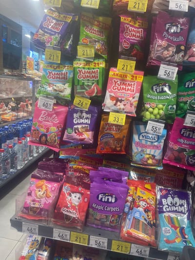 The image depicts a supermarket aisle in the city of Гожули, Ukraine, showcasing a variety of candy products. The shelves are densely packed with colorful bags and packages of gummies, jelly candies, and fruit snacks. Prominently displayed are brands like "YUMM! GUMMI," "Fini," "Barbie," "BOOM! GUMMI," "VIDAL," and "GUMMIZ" featuring characters from Paw Patrol. Prices are visible on some of the products, indicating a retail environment. The lighting suggests an indoor setting with artificial light sources. There are no people visible in the frame. The background shows more shelves with products, and to the left, bottled water is stacked on a lower shelf. The overall scene is a typical retail display of confectionery.