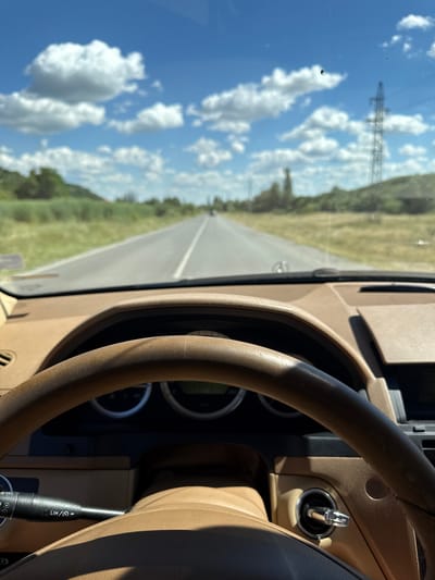 The image is a first-person perspective from inside a car, looking forward through the windshield. The car's interior is visible in the foreground, with a brown leather steering wheel and dashboard. A road stretches out ahead, with green fields on either side. The sky is blue with fluffy white clouds. In the distance, a power line tower can be seen on the right. The scene appears to be a rural landscape. The image was captured on a sunny day.