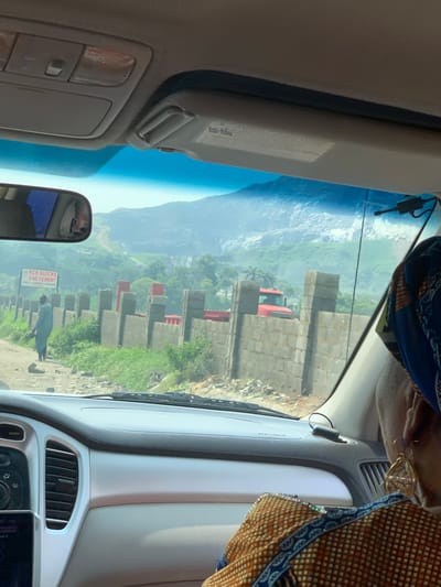 The image captures a view from inside a vehicle, looking out through the windshield and a side window. The scene appears to be a roadside in Dam Camp, Nigeria, with a construction site visible. Stacks of concrete blocks and some red building materials are piled along the road. A person, dressed in a light blue garment, is walking on the right side of the road. Green vegetation and a hazy, mountainous landscape form the background. The sky is bright blue, suggesting it is daytime, likely late morning or early afternoon, with clear weather. Inside the car, part of the dashboard and a sun visor are visible. A person's head, wearing a patterned headwrap and jewelry, is partially visible on the right side of the frame, suggesting the photographer is a passenger. A rearview mirror shows a reflection of the interior of the car. A sign with text is visible by the road; it reads "RCR BLOCKS AND CEMENT". The overall impression is a typical scene of daily life and ongoing construction in a rural or semi-urban area of Nigeria.