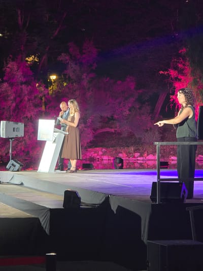 An outdoor nighttime event is taking place on a stage with pink and purple lighting. On the stage, a man in a suit and a woman in a brown dress are standing behind a podium with a large illuminated number "7" on it. The woman is holding papers and appears to be speaking. To the right of the stage, a woman with curly hair is standing and pointing to her left. The background features trees illuminated with pink light and a body of water. The event is located in Cornellà de Llobregat, Spain.