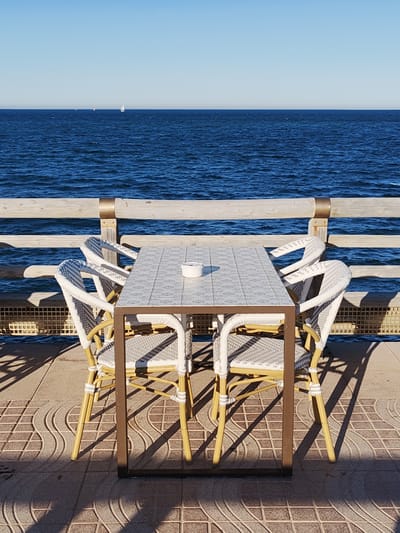 Here is a description of the image:
The image shows a dining table and four chairs on a tiled patio. The table has a tiled surface and a brown metal frame. The chairs are white with a woven pattern and yellow legs. On the table is a small, white, cylindrical container. The patio overlooks a wooden railing, beyond which is the sea. A sailboat is visible on the horizon. The scene is bathed in bright sunlight, casting shadows of the table and chairs onto the patio. The location is likely a seaside restaurant or cafe in Dénia, Spain. There are no people visible in the frame.